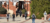 Students with backpacks, seen from behind, walking toward a red brick campus building with white pergola details on a sunny day.