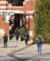 Students with backpacks, seen from behind, walking toward a red brick campus building with white pergola details on a sunny day.