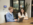 Two professionals collaborating at a wooden conference table in a modern office setting. Joey, an associate marketing strategist, is in a light blue quarter-zip pullover and, Alex, an account executive, is in a cream-colored lace top are both smiling and looking at a laptop screen together. The office features floor-to-ceiling windows, a large plant, and contemporary furniture. There's a tablet on the table and a digital display mounted on the wall in the background.